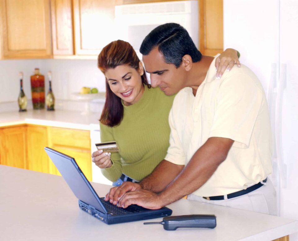 A couple stands in the kitchen using their credit card to purchase something over the internet